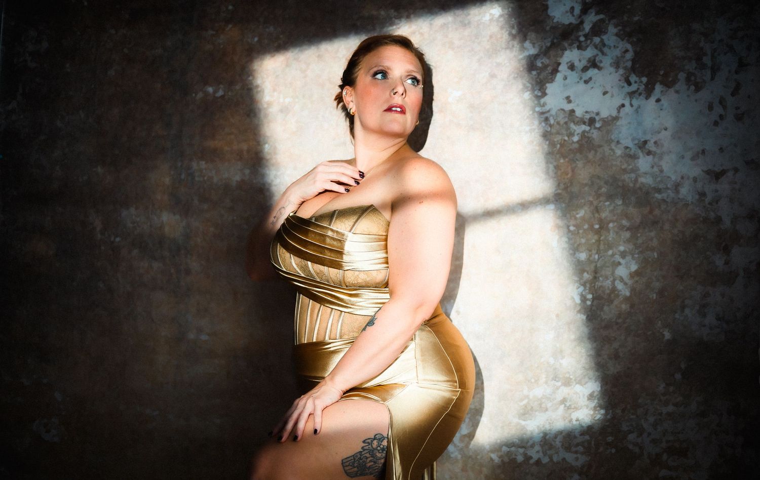 A person poses in a shimmering gold dress against a moody concrete wall with dramatic lighting and shadows.