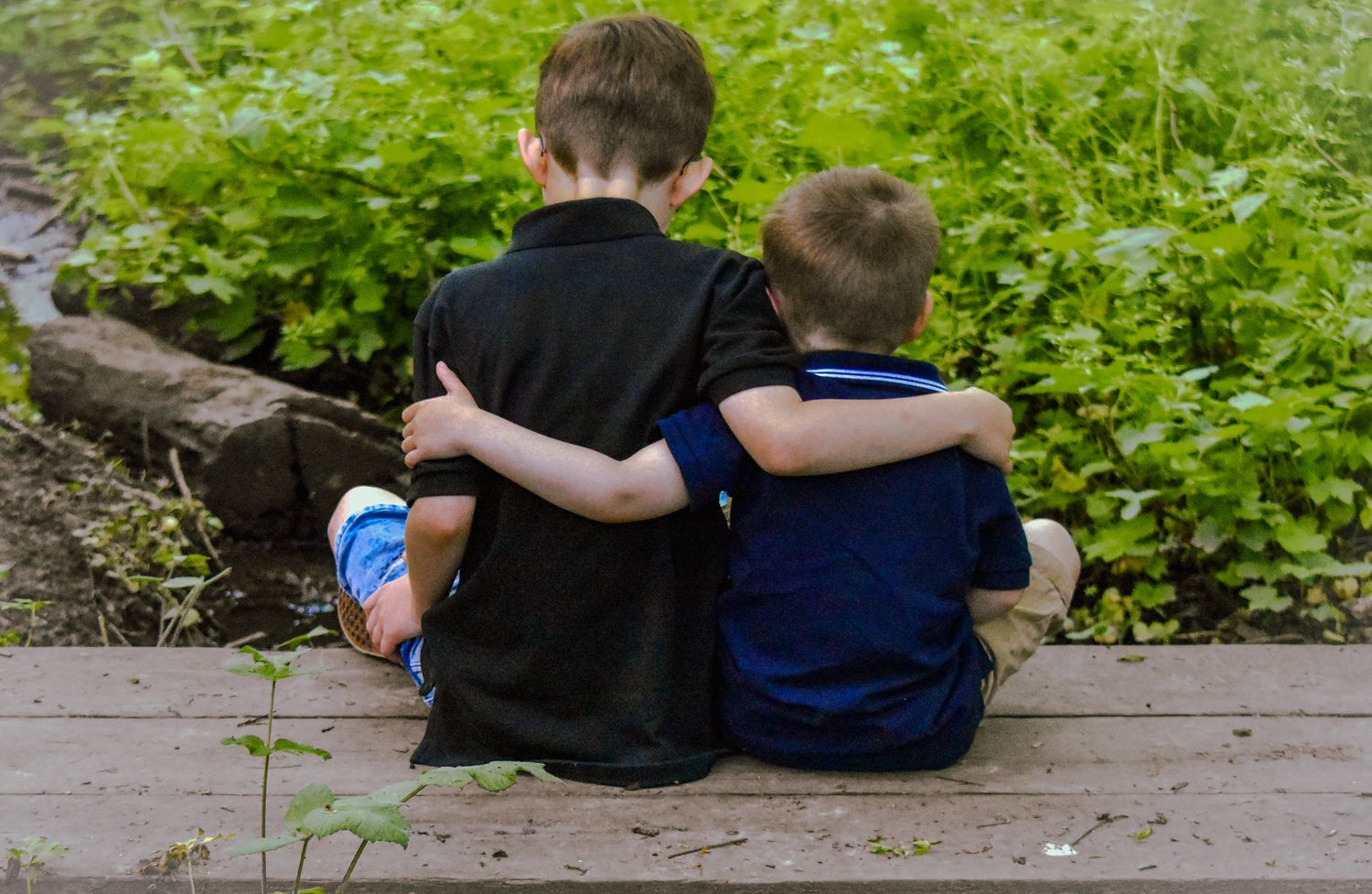 Two children sitting together on wooden deck surrounded by greenery.