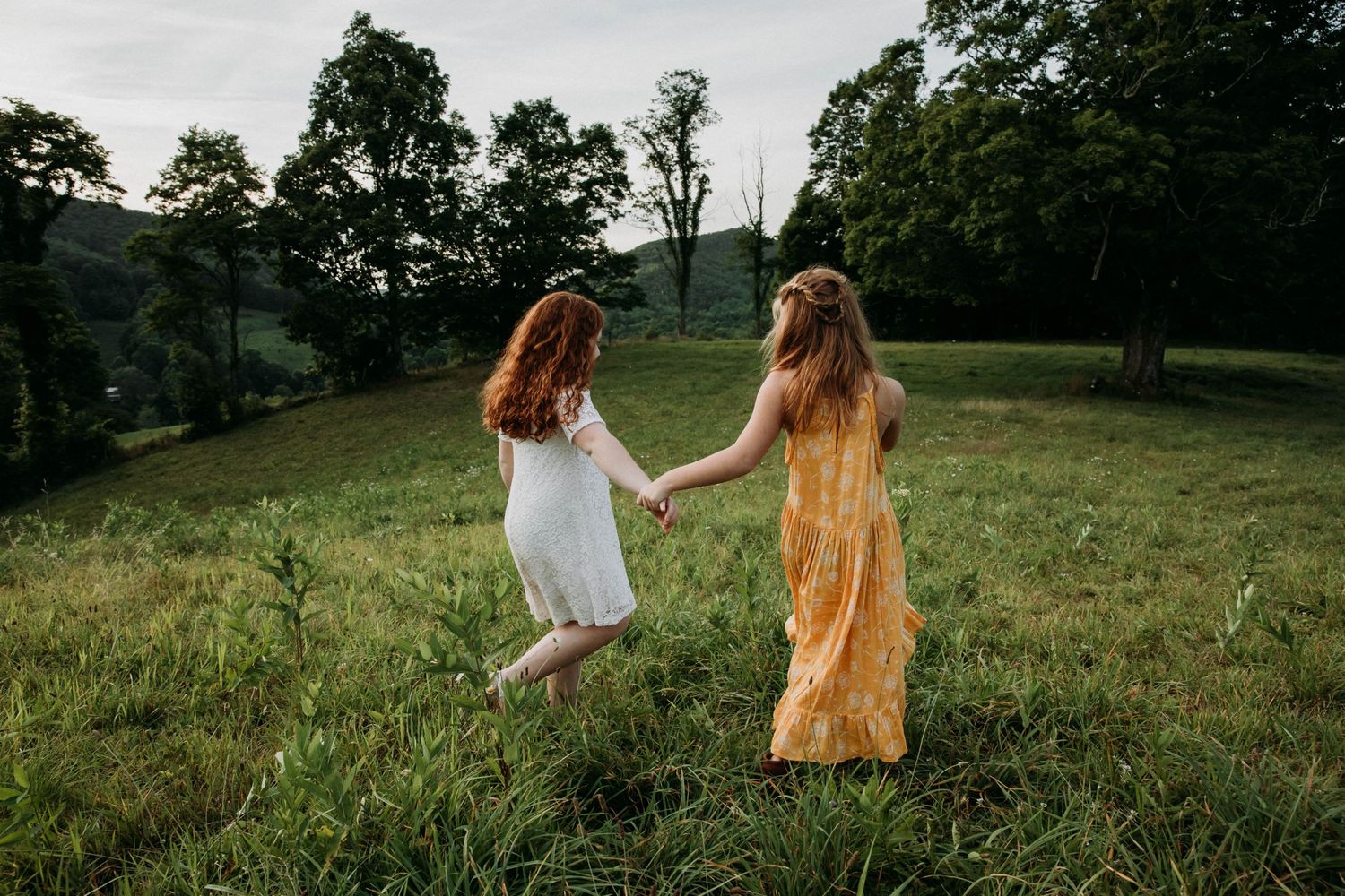 Photo of a girl in a white dress and a girl in a yellow dress walking through a field holding hands in Spring