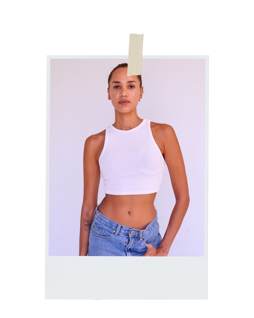 Athletic figure in a white cropped tank top and high-waisted blue jeans poses against a light purple background.