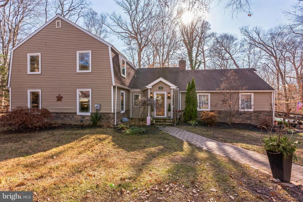 Tan colonial-style home with brick accents features gambrel roof and central entrance walkway in wooded setting.