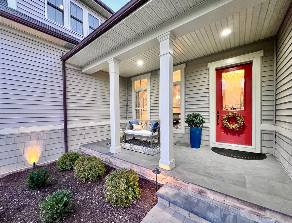 Cozy front porch with white columns, a bright red door with wreath, and comfortable seating against gray siding.