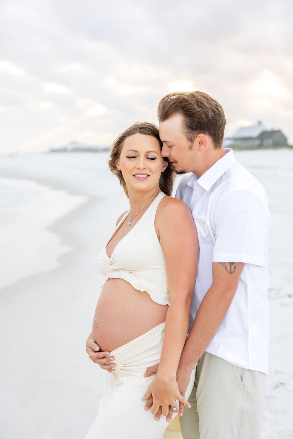 A couple shares an intimate moment on a white sandy beach with the ocean and cloudy sky in the background.