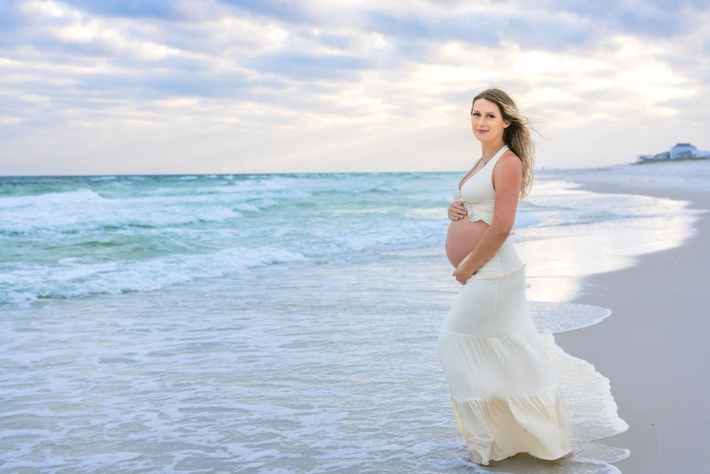 Pregnant woman in white flowing dress stands on beach as ocean waves roll in under cloudy sky.