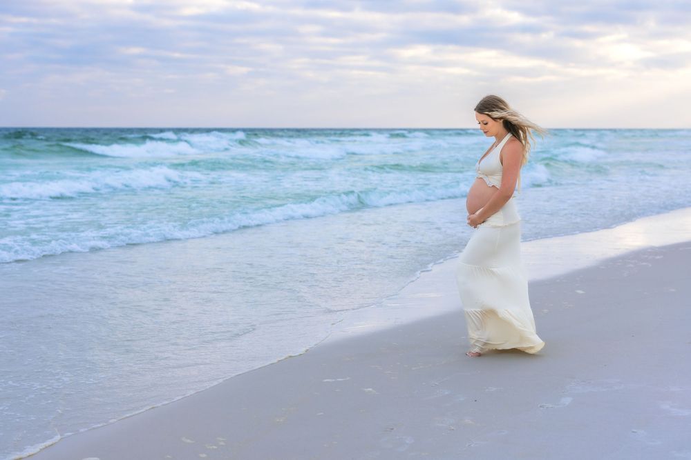 A figure in a flowing white dress walks along a beach at sunset while waves crash against the shore.