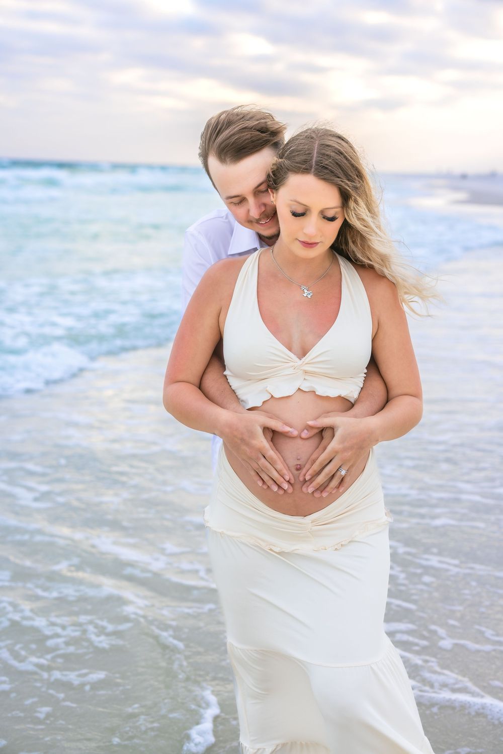 A romantic maternity photo of a couple embracing on the beach at sunset with waves in the background.