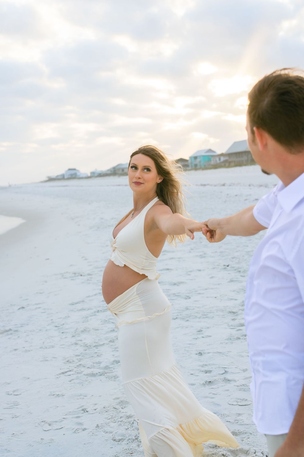 Couple dances together on a white sandy beach with coastal homes visible in the background at sunset.