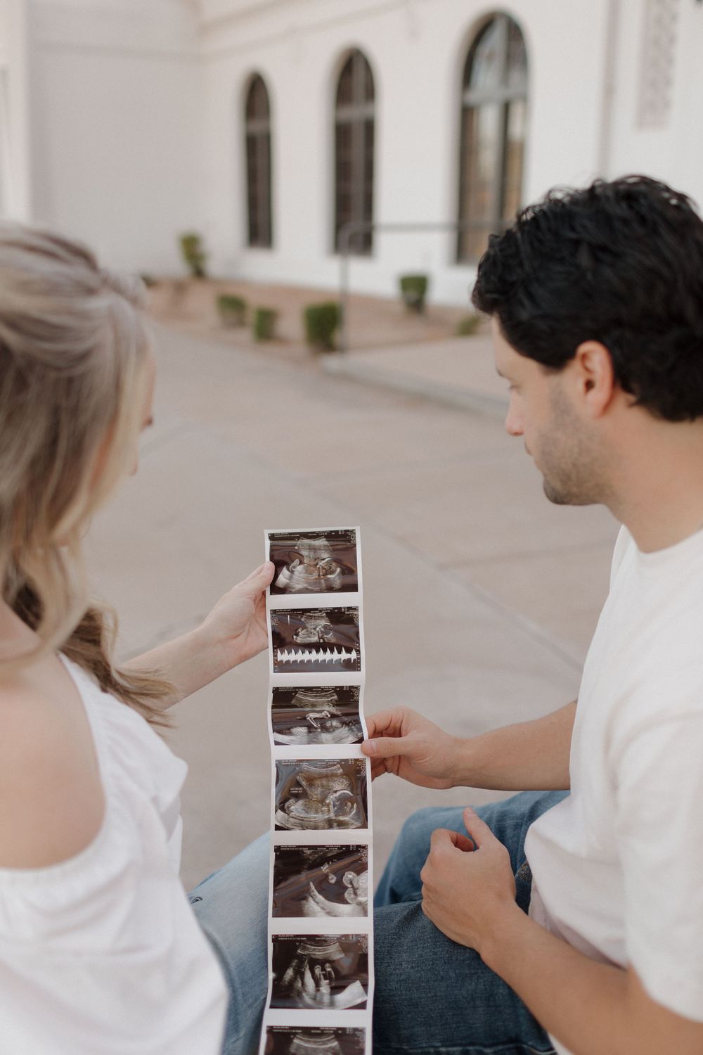Couple examining ultrasound photos together while sitting outside white building.