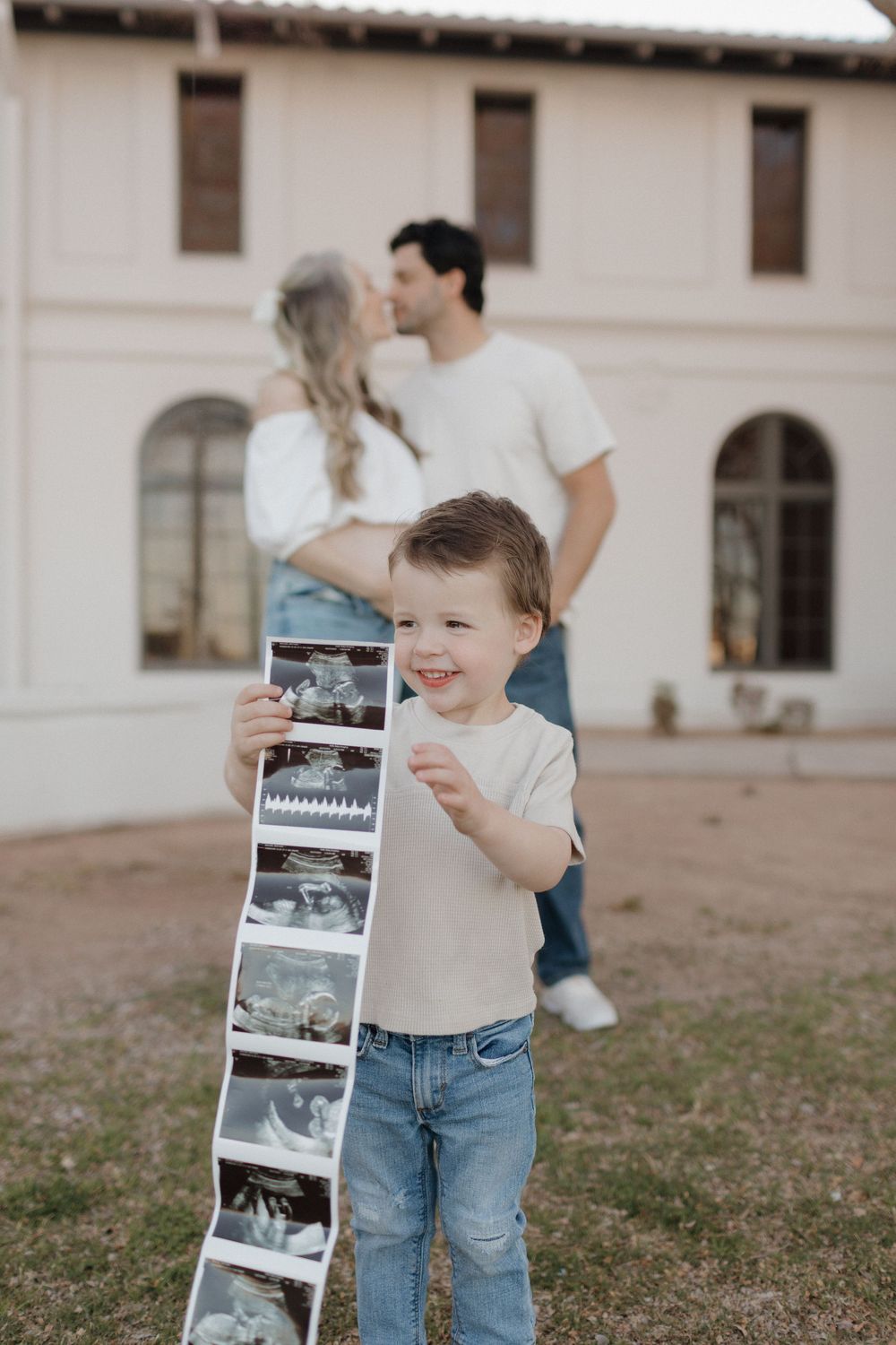 A young child examines ultrasound photos with curiosity outside a white building.
