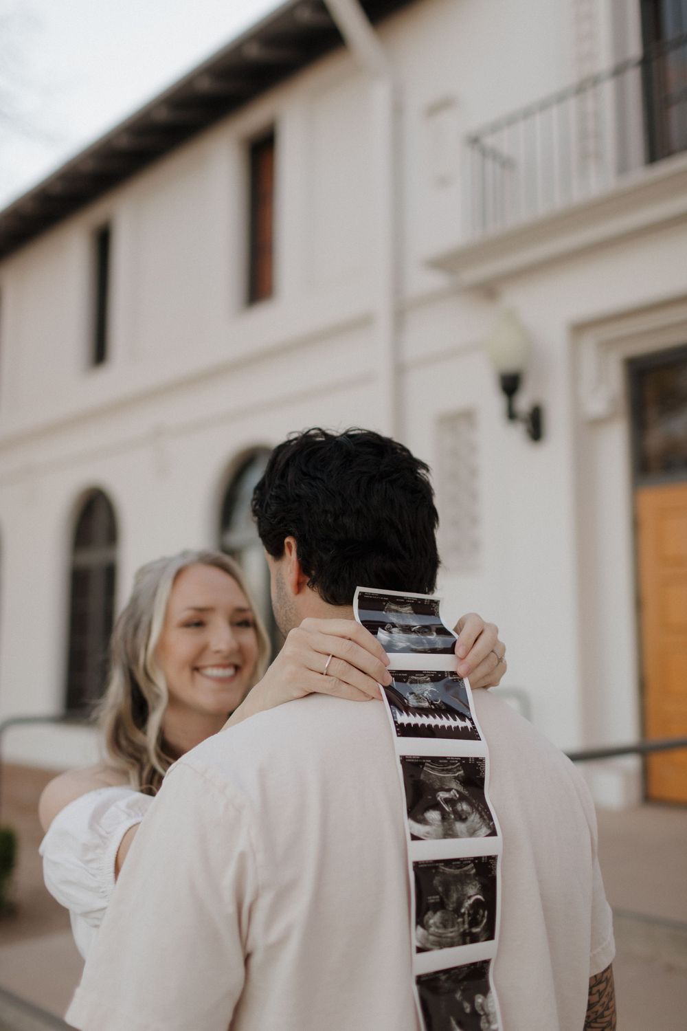 Joyful couple holding ultrasound photos outside a white building during pregnancy announcement.