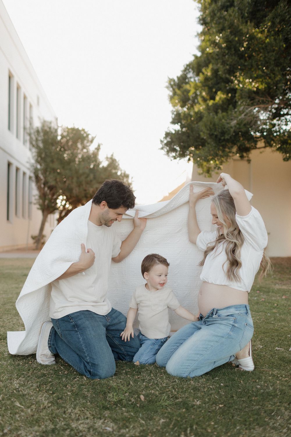 Happy family plays peek-a-boo with white fabric during outdoor photoshoot.