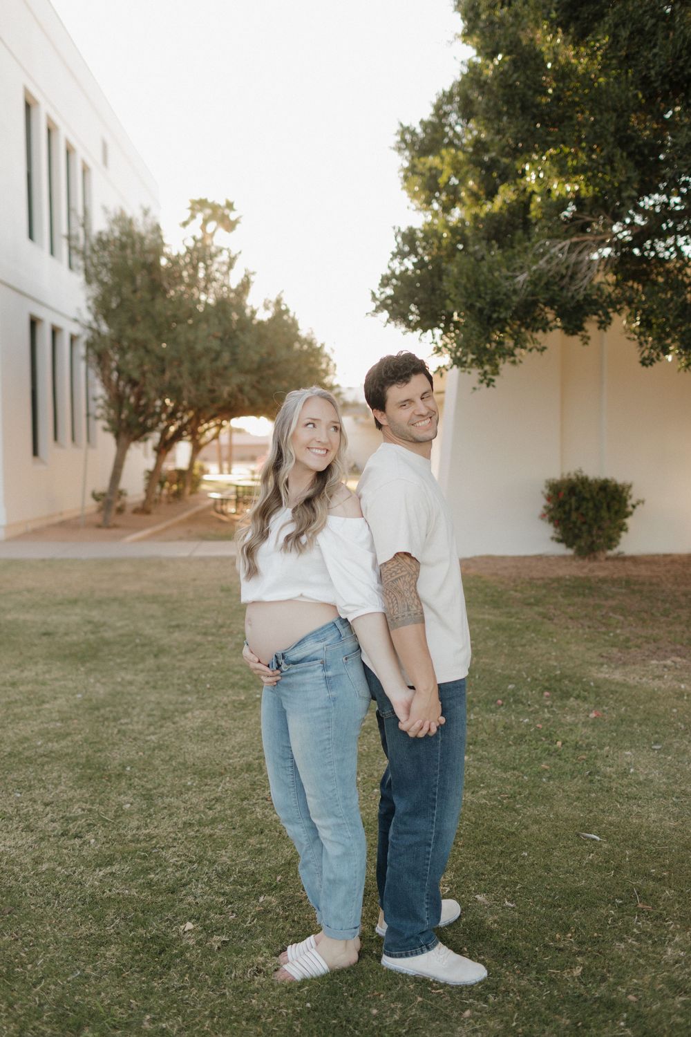 Expectant couple in casual attire standing together on grass at sunset.