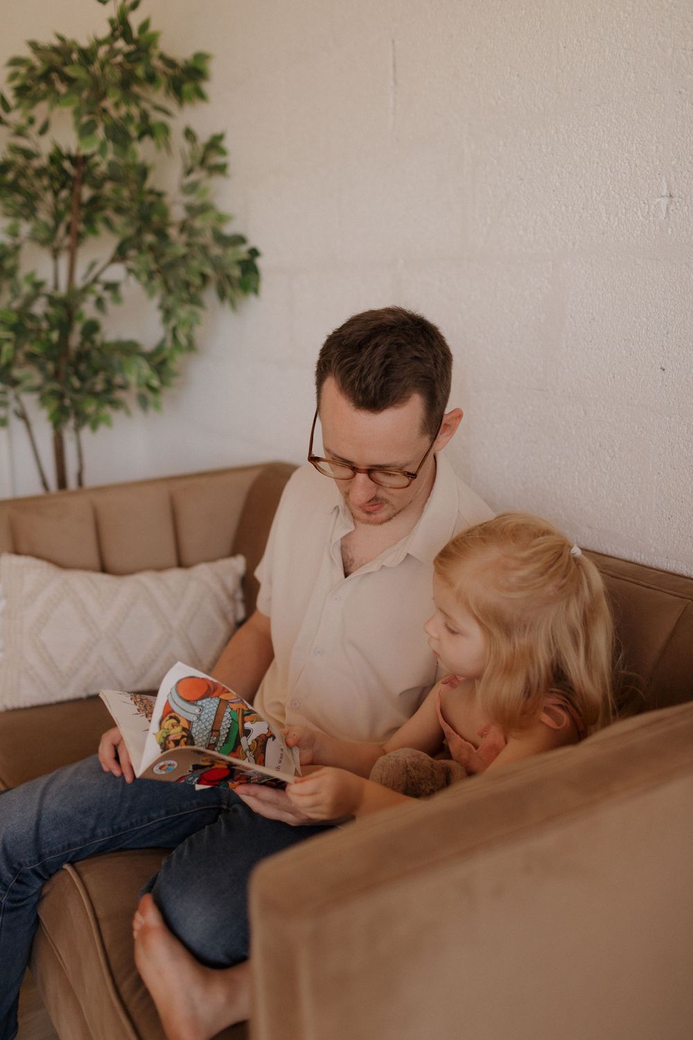 Cozy living room scene with figures sharing a reading moment on a brown couch.