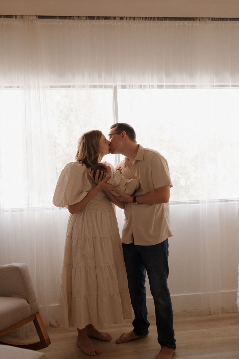 Couple shares a kiss while holding their newborn baby in a softly lit nursery setting.