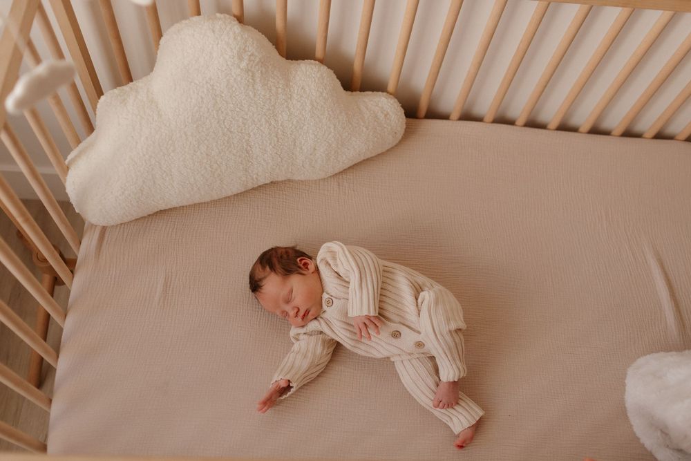 A sleeping baby in white clothing rests peacefully in a wooden crib with cloud pillow.