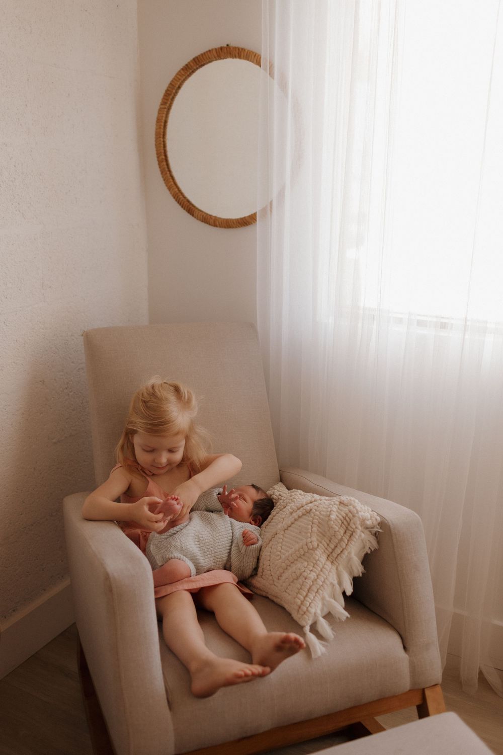 A sunlit room shows a child and baby sharing quiet moments on a grey couch.