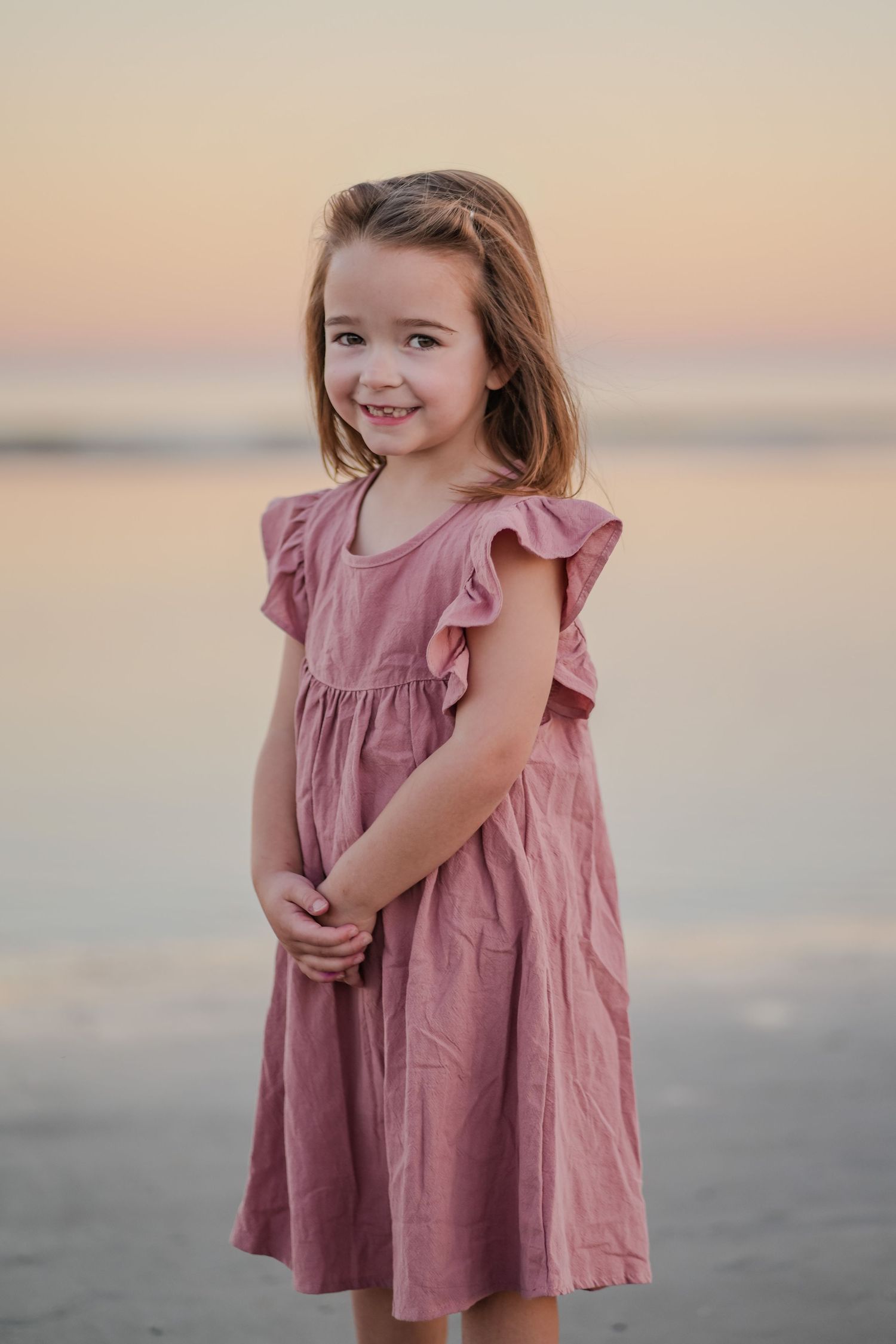 Smiling young girl in a pink ruffle dress standing on a beach at sunset.
