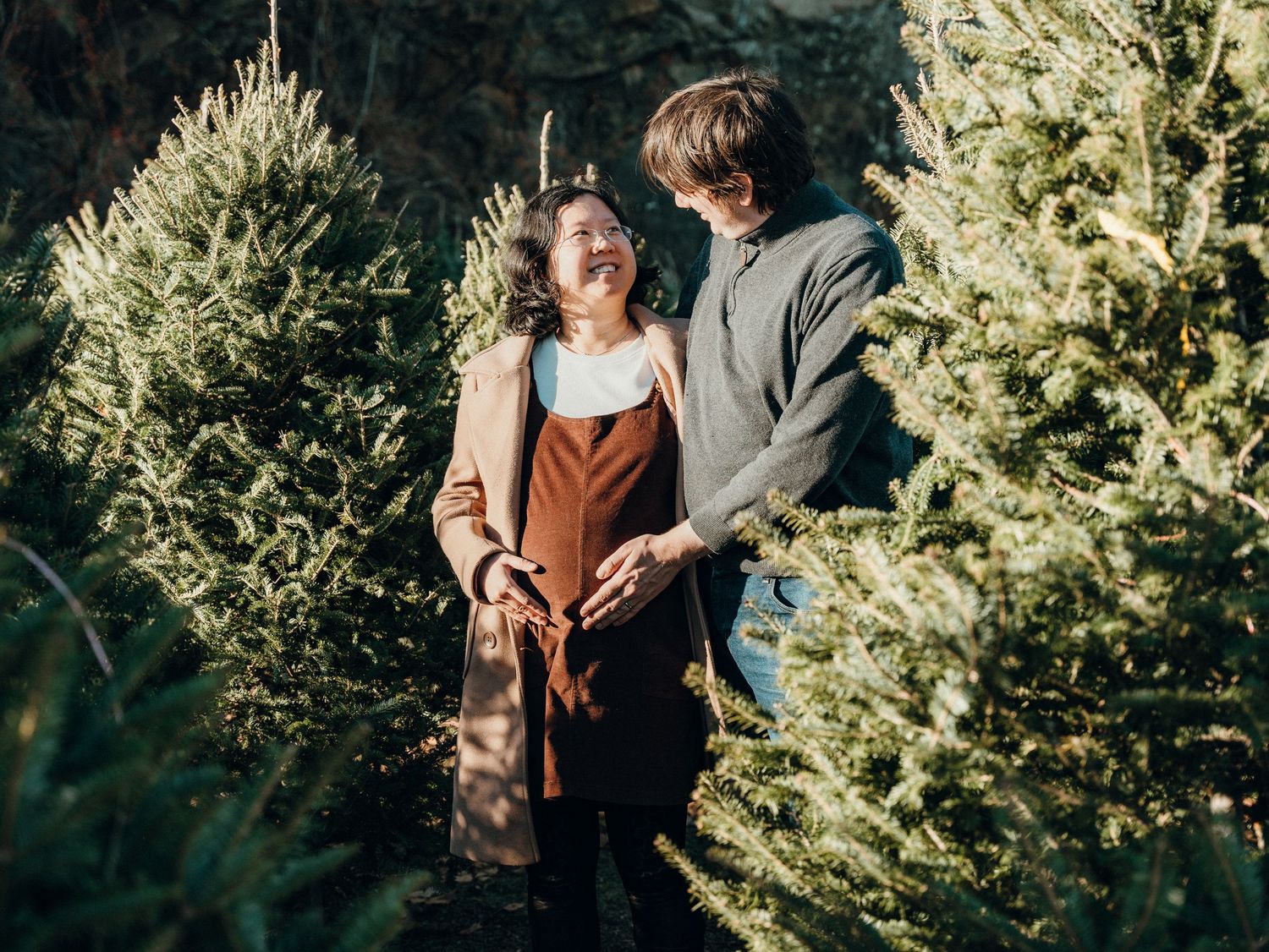 A couple poses among evergreen trees during a winter Christmas theme photoshoot.