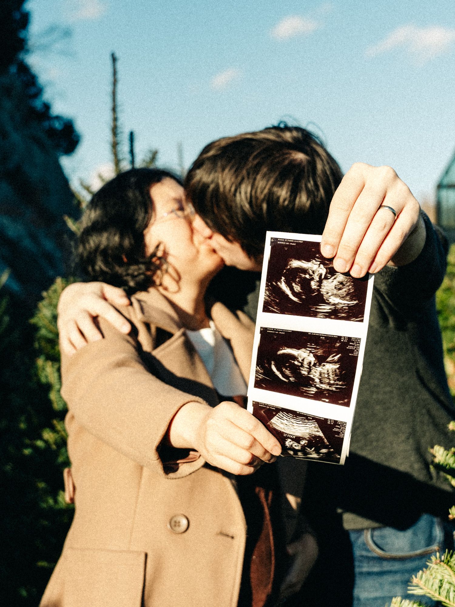 A couple holds up ultrasound photos while sharing an intimate moment outdoors.