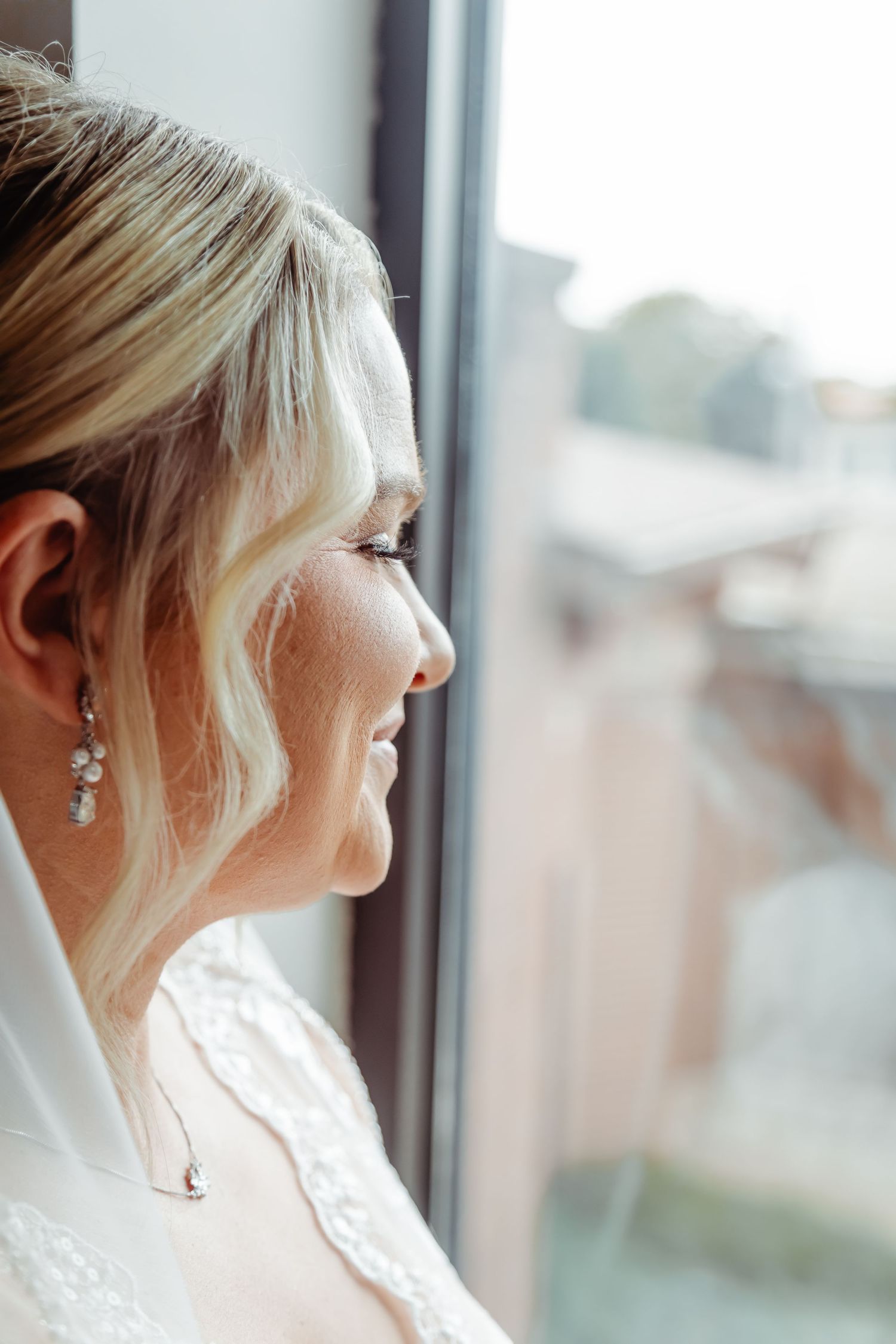 Profile view of a bride in a white dress with blonde hair and crystal earrings gazing out a window.