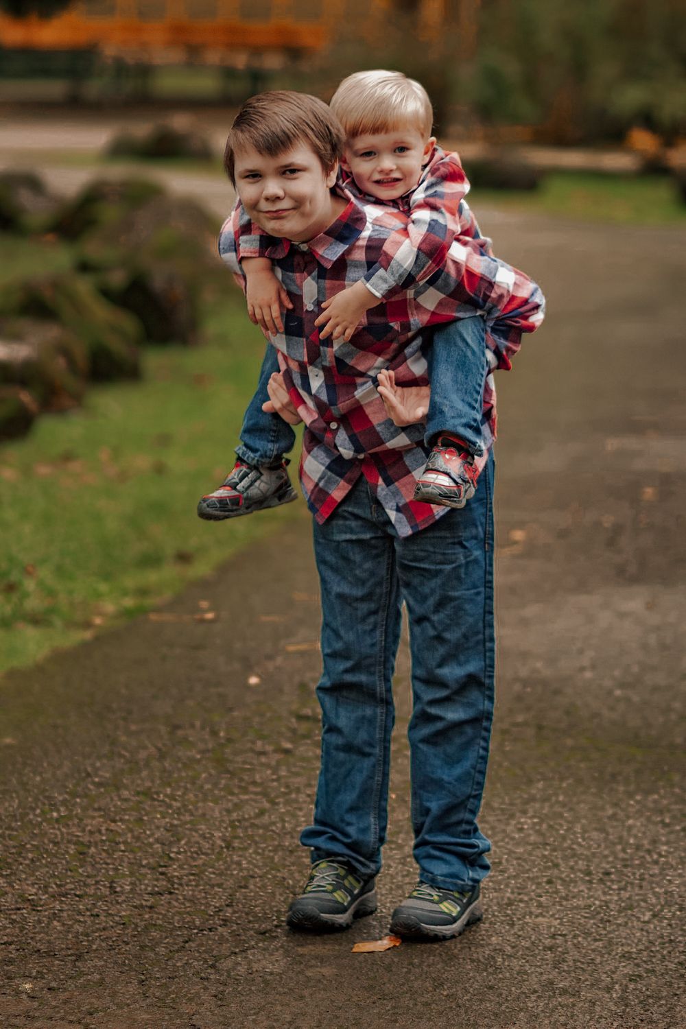 Older brother giving younger sibling a piggyback ride on a park path in matching plaid shirts.