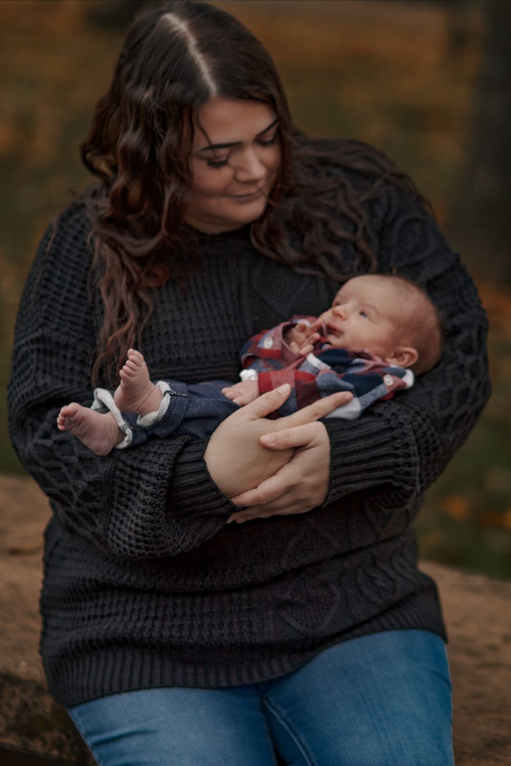 Mother in dark knit sweater lovingly gazing at her newborn baby in an autumn park setting.