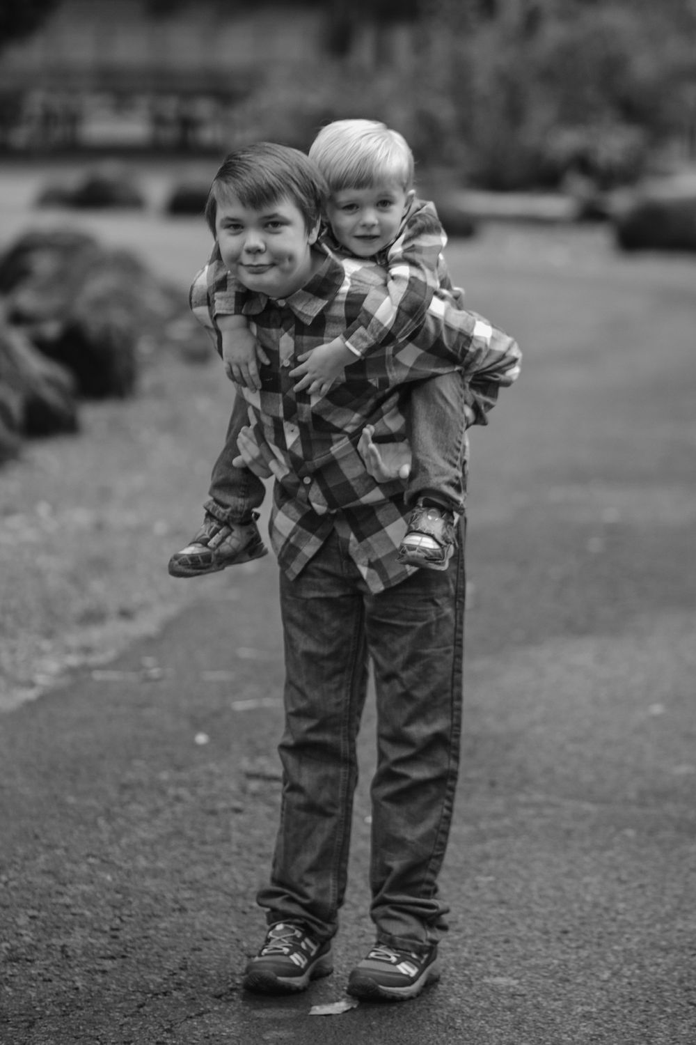Black and white photo of older boy giving younger sibling a piggyback ride outdoors.
