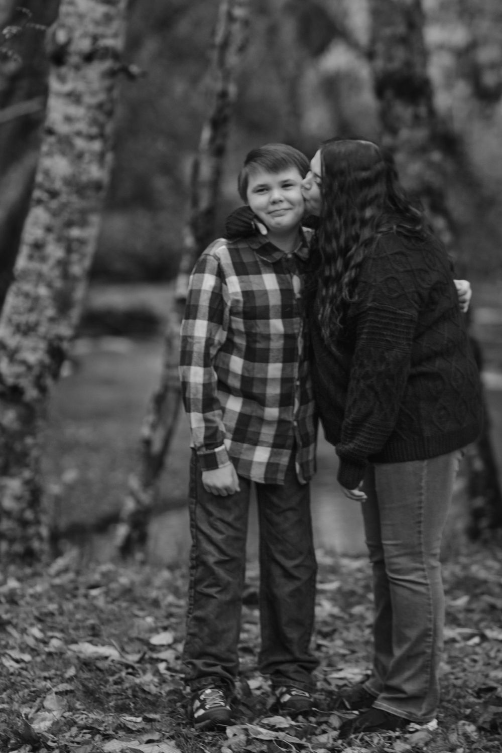 Black and white photo of mother kissing smiling boy on cheek among autumn trees.