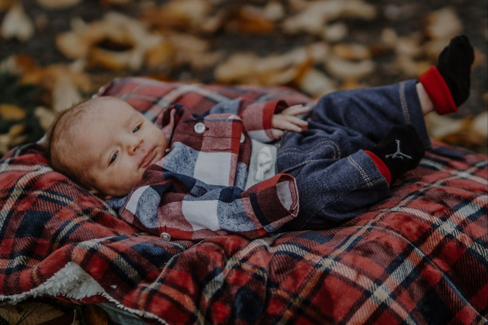 Newborn baby in red plaid shirt lying on a matching plaid blanket surrounded by autumn leaves.