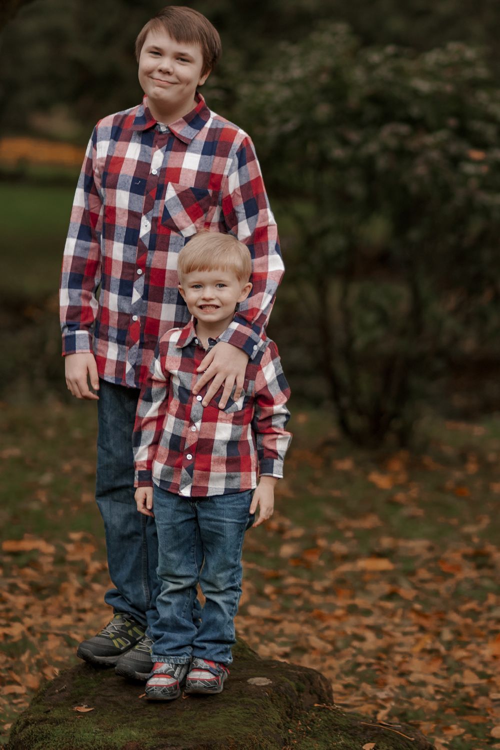 Two brothers in matching red plaid shirts posing on a mossy rock in an autumn park.