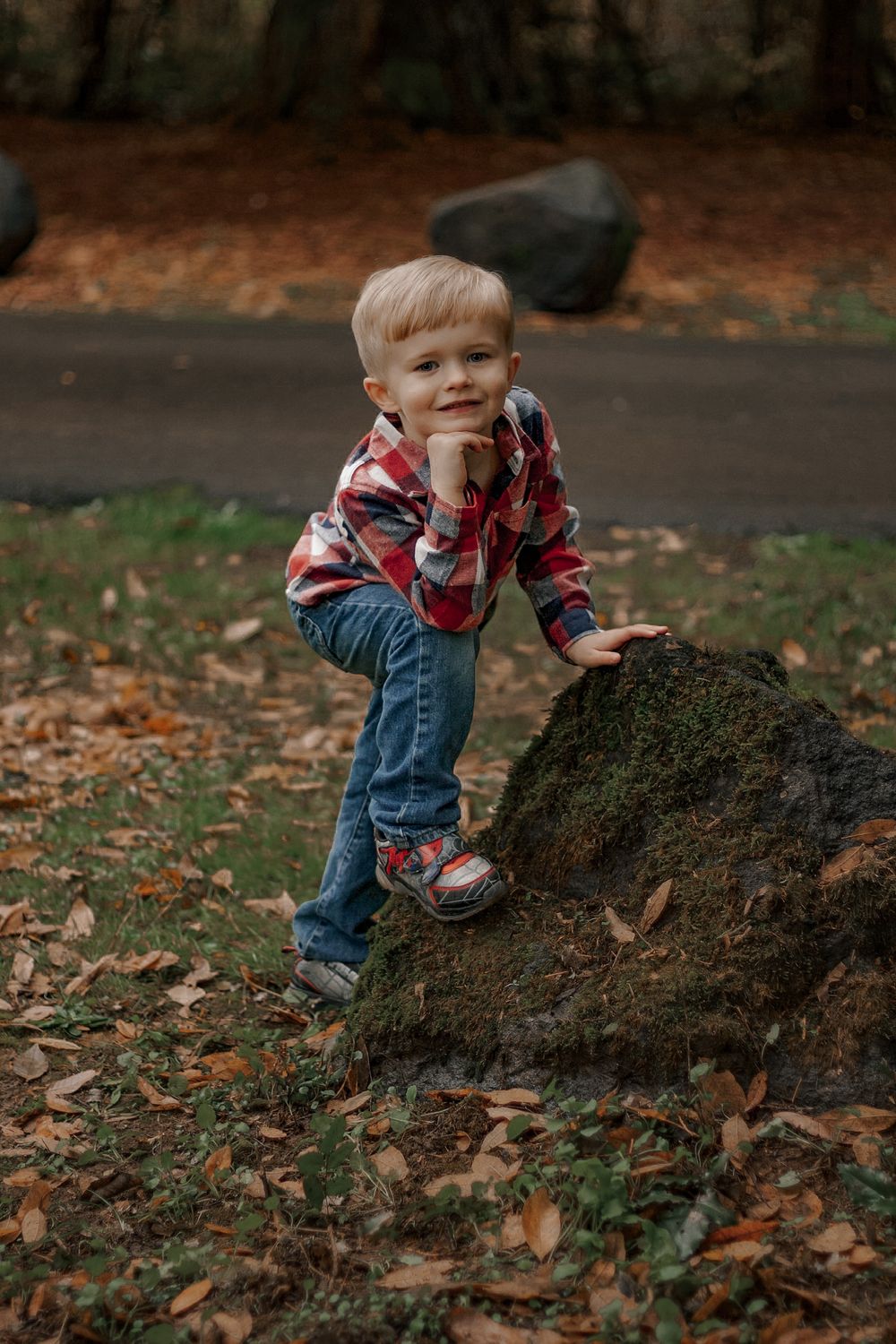 Adorable blonde toddler in red plaid shirt climbing a mossy rock in an autumn park.