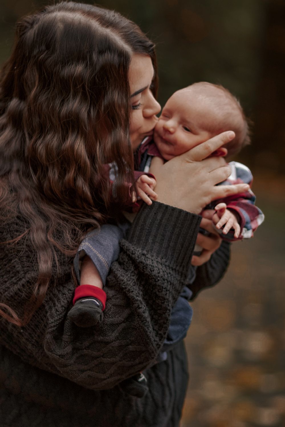 Mother tenderly kissing and holding smiling newborn baby outdoors in autumn setting.