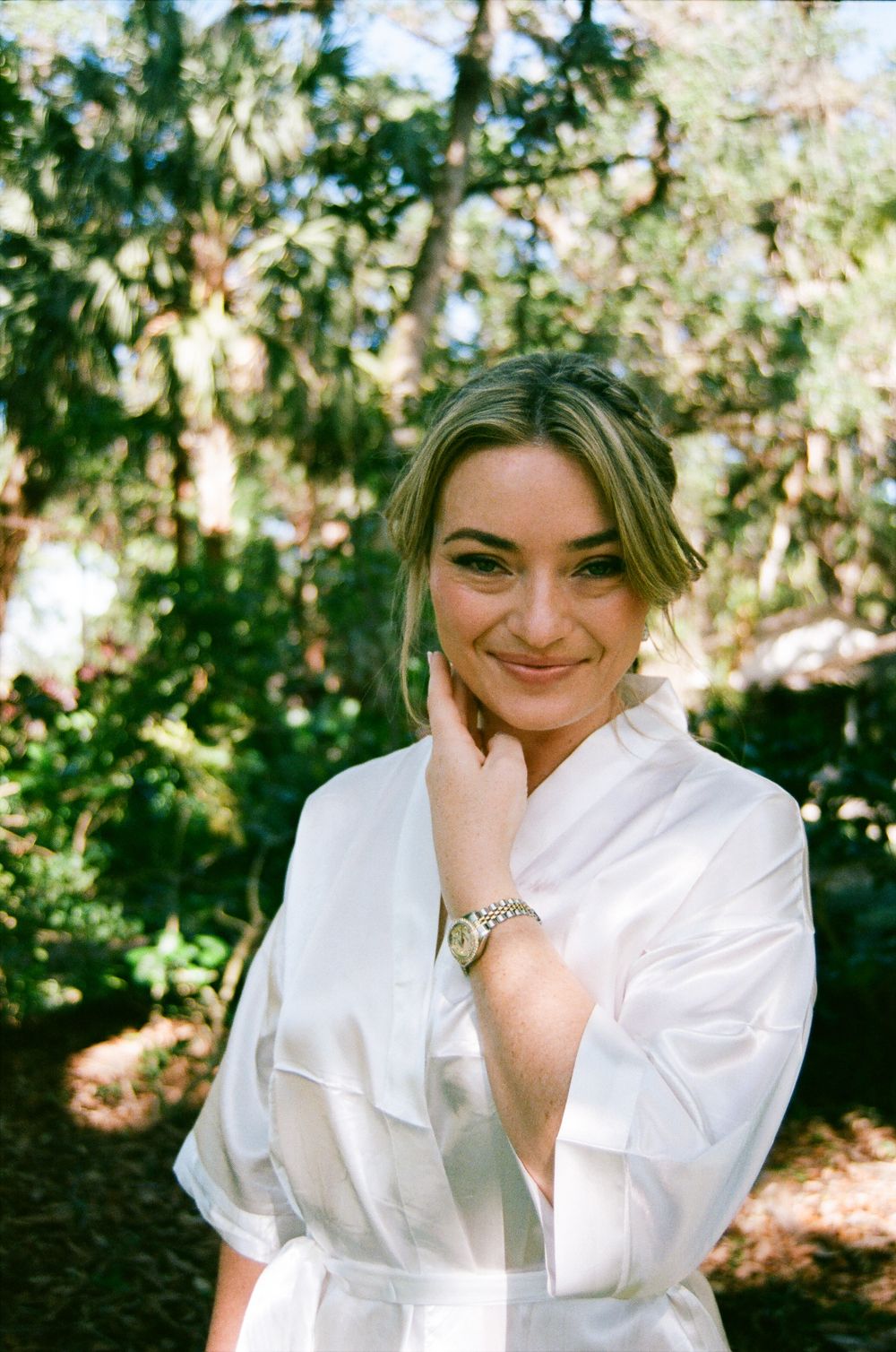 A smiling woman in a white satin robe wearing a watch poses outdoors among lush green trees.