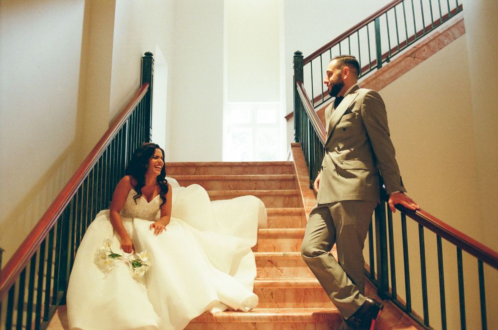Bride in white gown and groom in gray suit share a romantic moment on elegant staircase during wedding day.