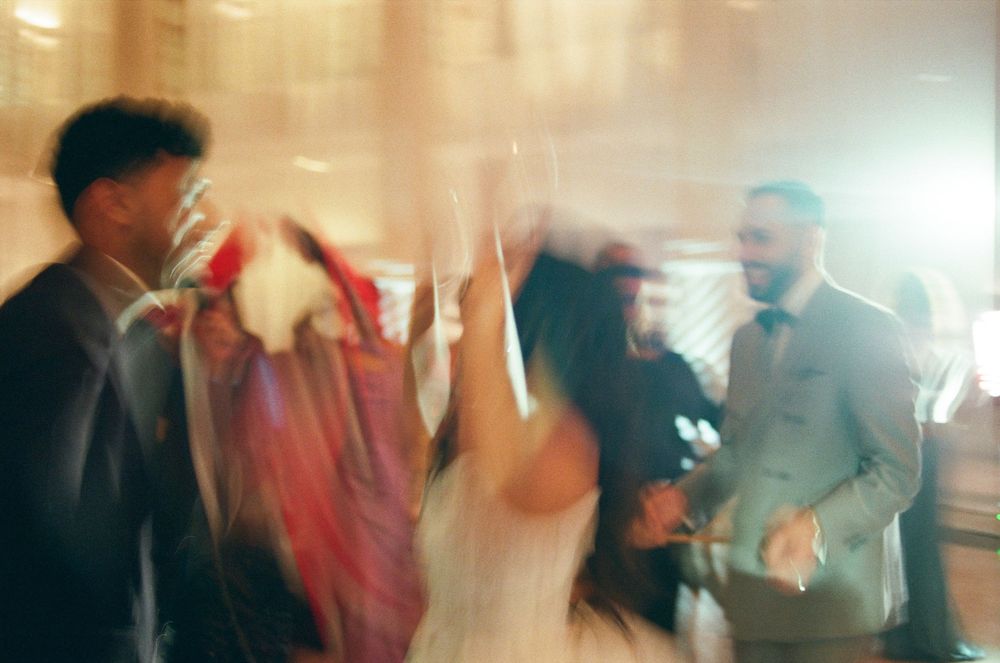 Blurry motion shot of wedding guests dancing joyfully at a reception, bride in white gown surrounded by celebrants.