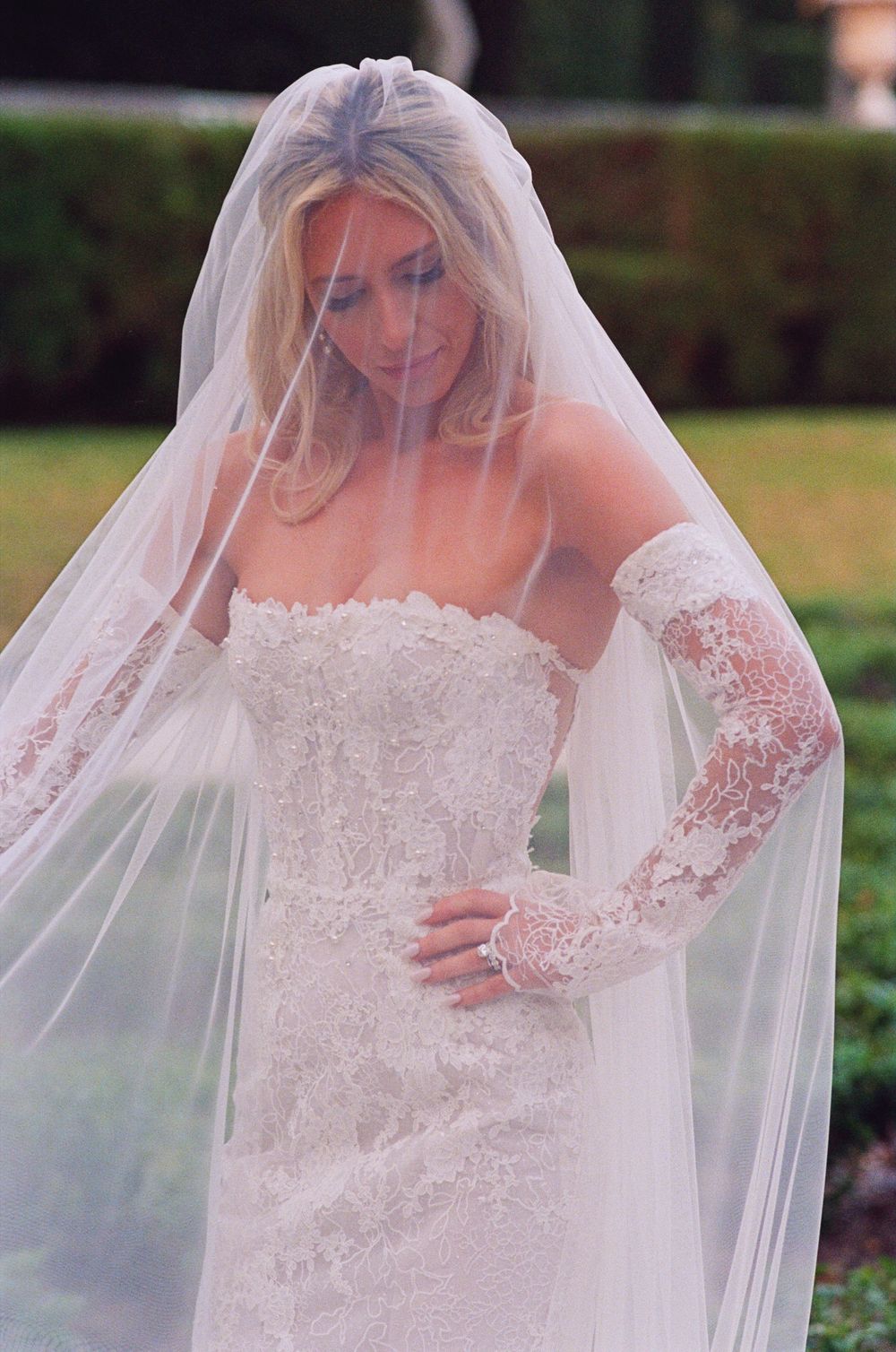 A bride poses in an elegant strapless lace wedding gown featuring intricate long sleeves and a flowing veil outdoors.