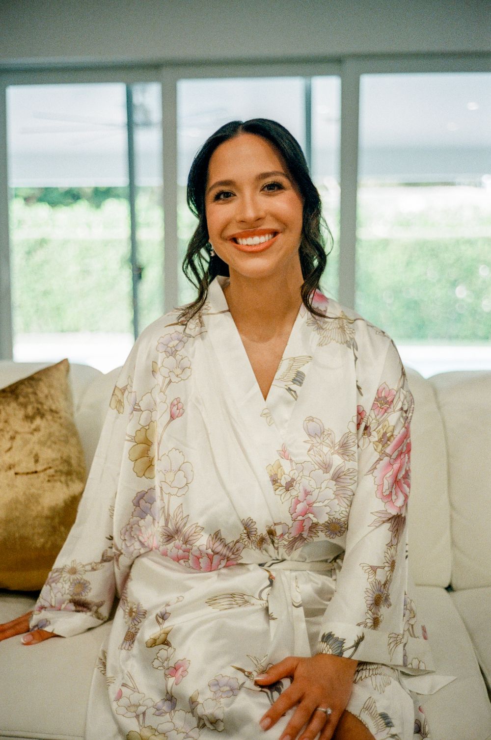A person wearing a white floral robe sits on a couch smiling during wedding day preparations.