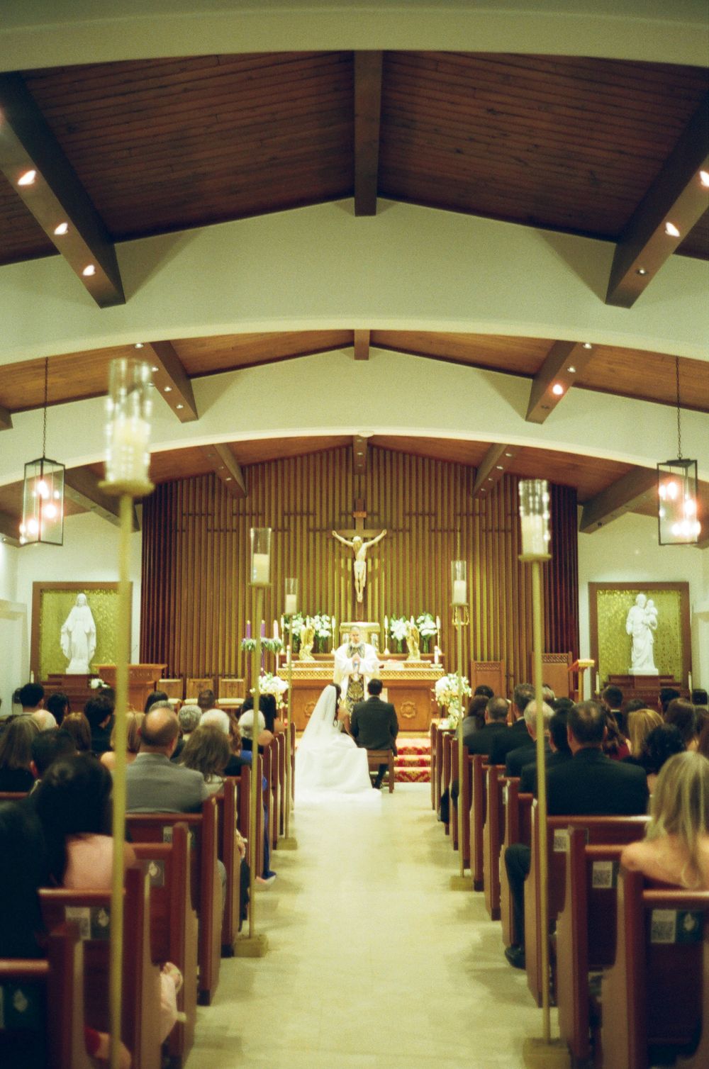 Interior view of a Catholic church during a wedding ceremony with wooden pews, vaulted ceiling and hanging lanterns.