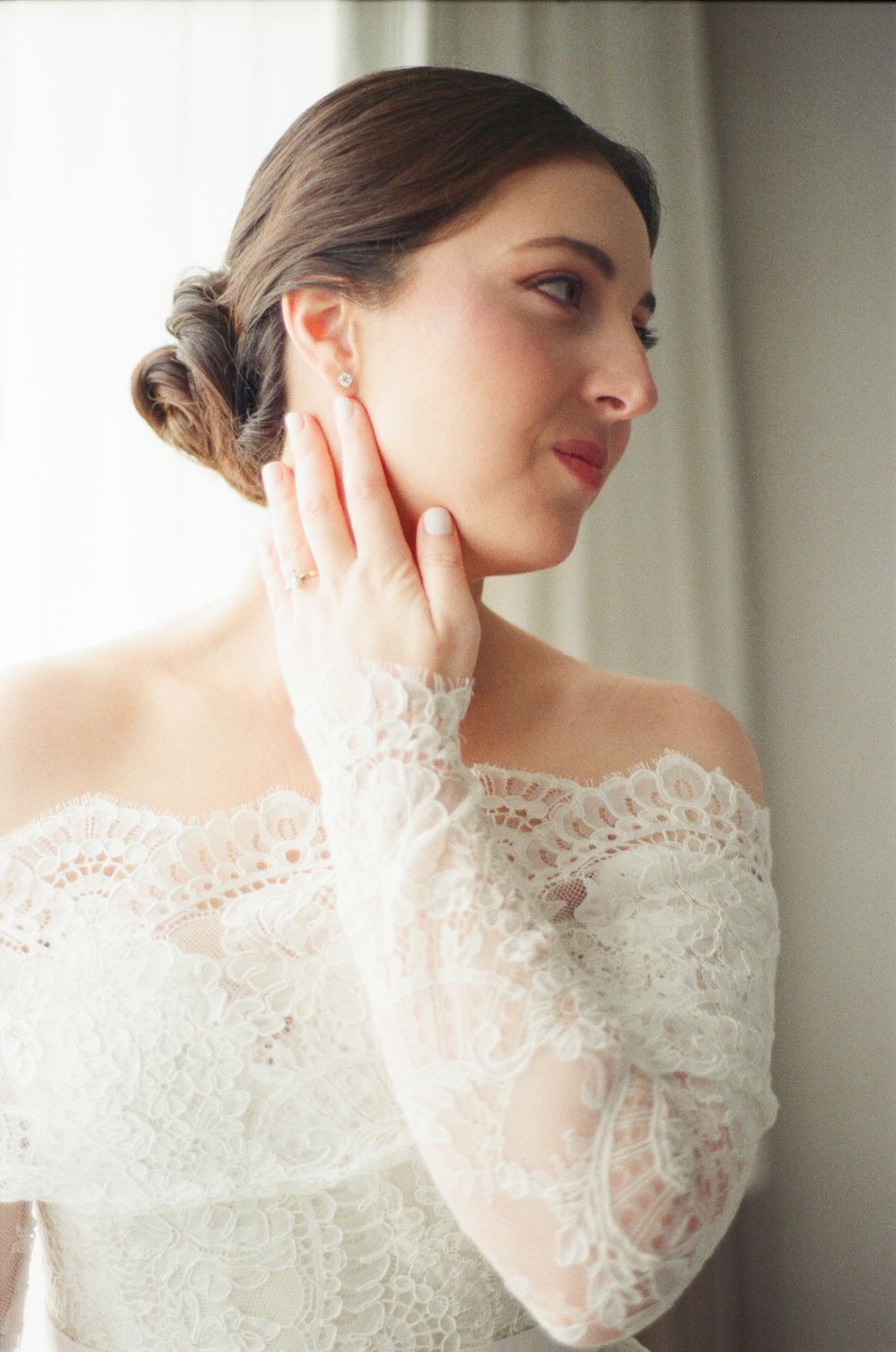 Bride in off-shoulder lace wedding dress with elegant updo, gently touching her face near a window.