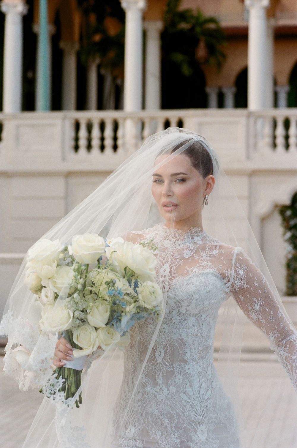 Bride in elegant lace long sleeve wedding gown holds luxurious white rose bouquet against classic columned backdrop.