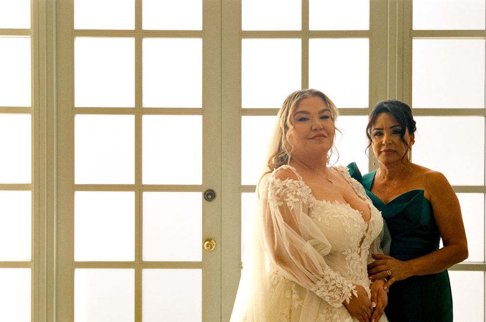 A bride in a white lace gown stands in front of French doors during wedding preparations.