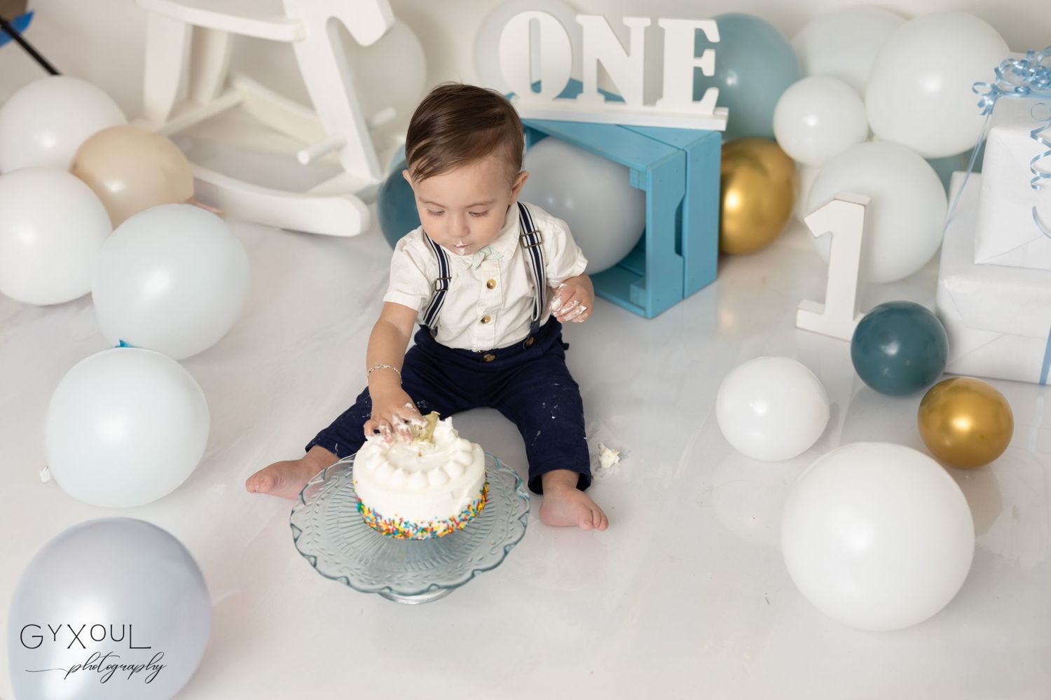 Baby's first birthday cake smash setup with blue and gold balloons and a rocking horse decoration.