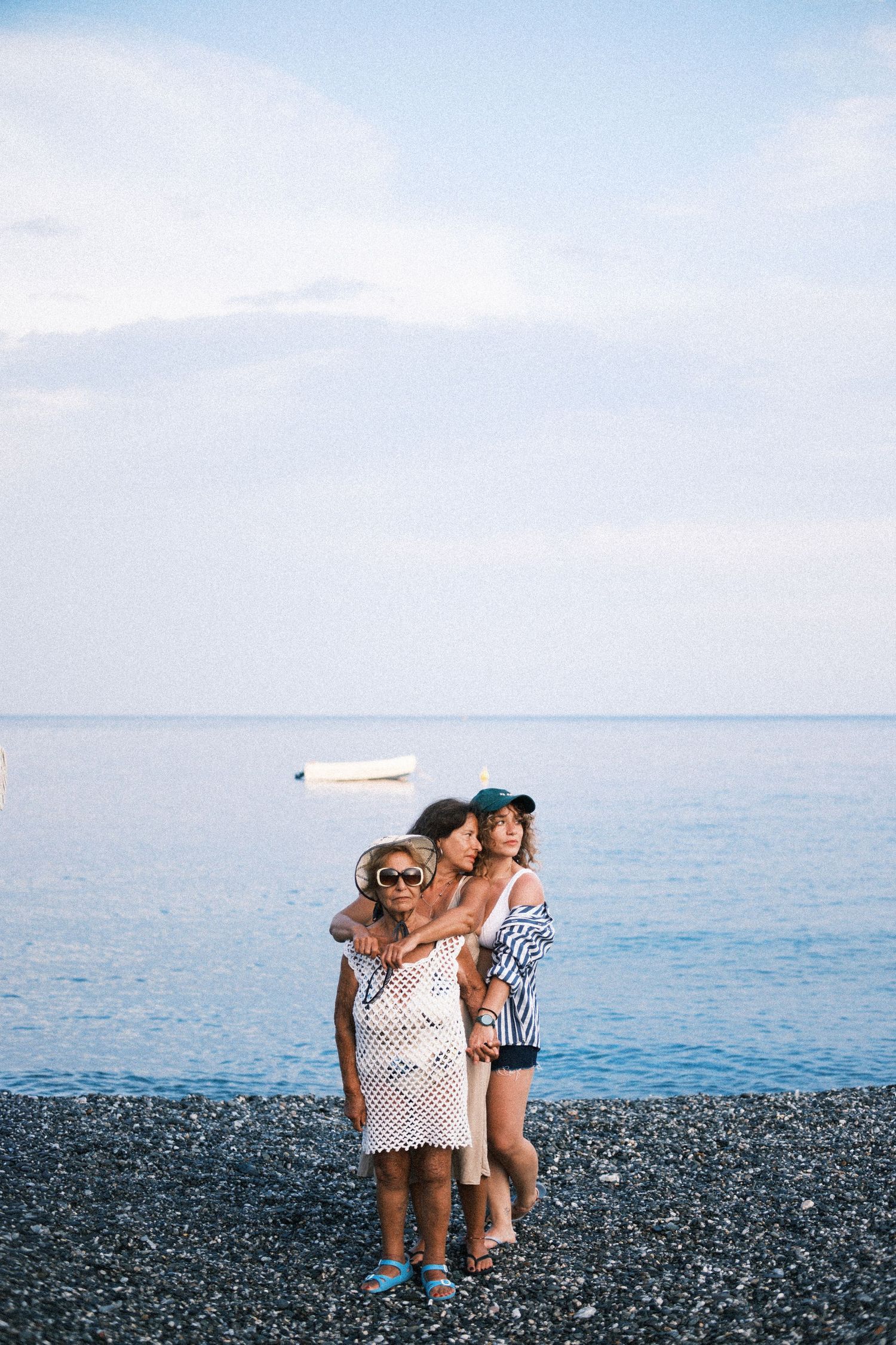 Three people enjoying a sunny day together on a rocky beach with calm blue waters and clear skies in the background.