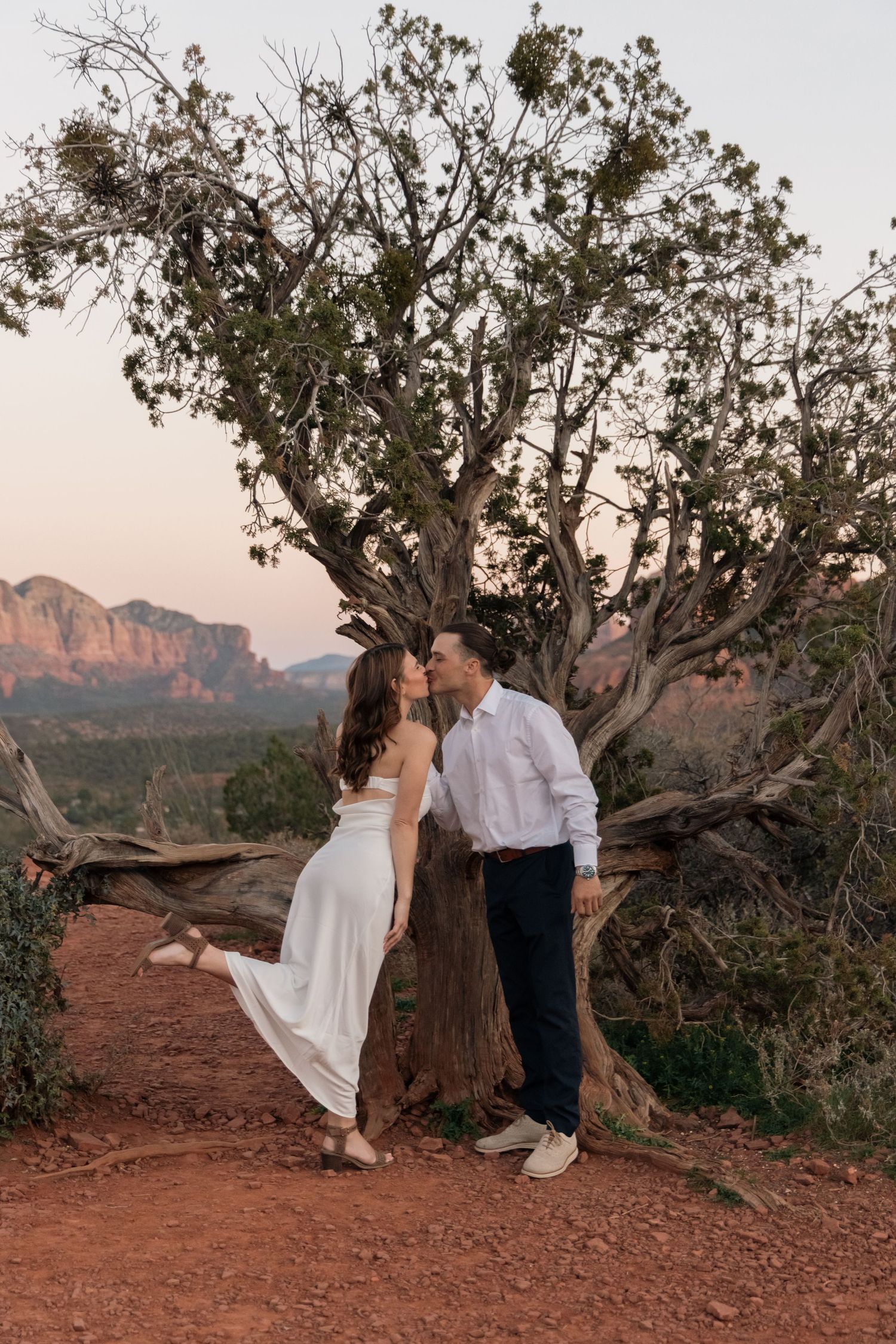 A romantic couple poses together on red rocks during sunset in Sedona with mountains and desert landscape in background.