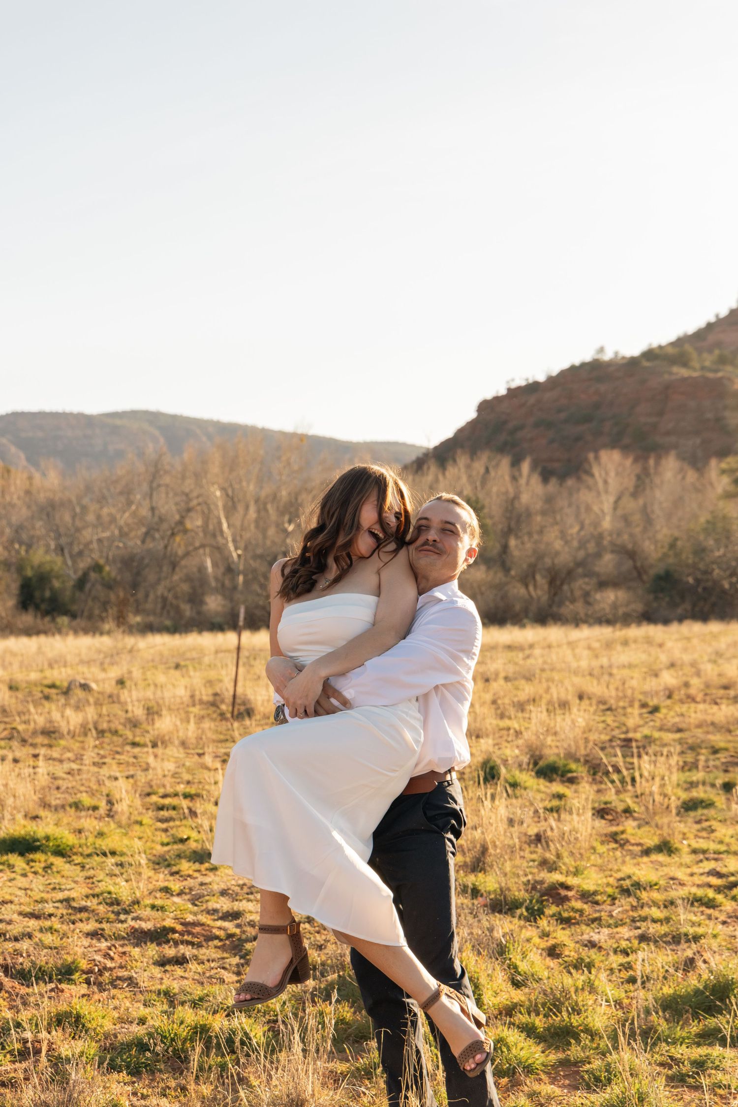 A couple in white embraces in a golden field at sunset with mountains in the background.