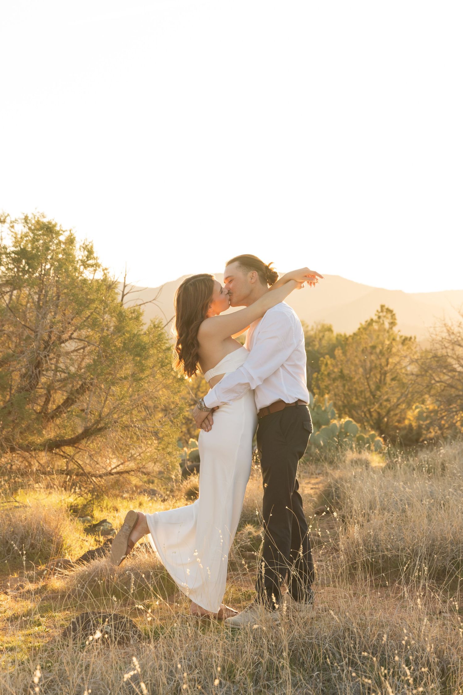 A romantic couple embraces in a golden sunset on a hillside with mountains and trees in the background.