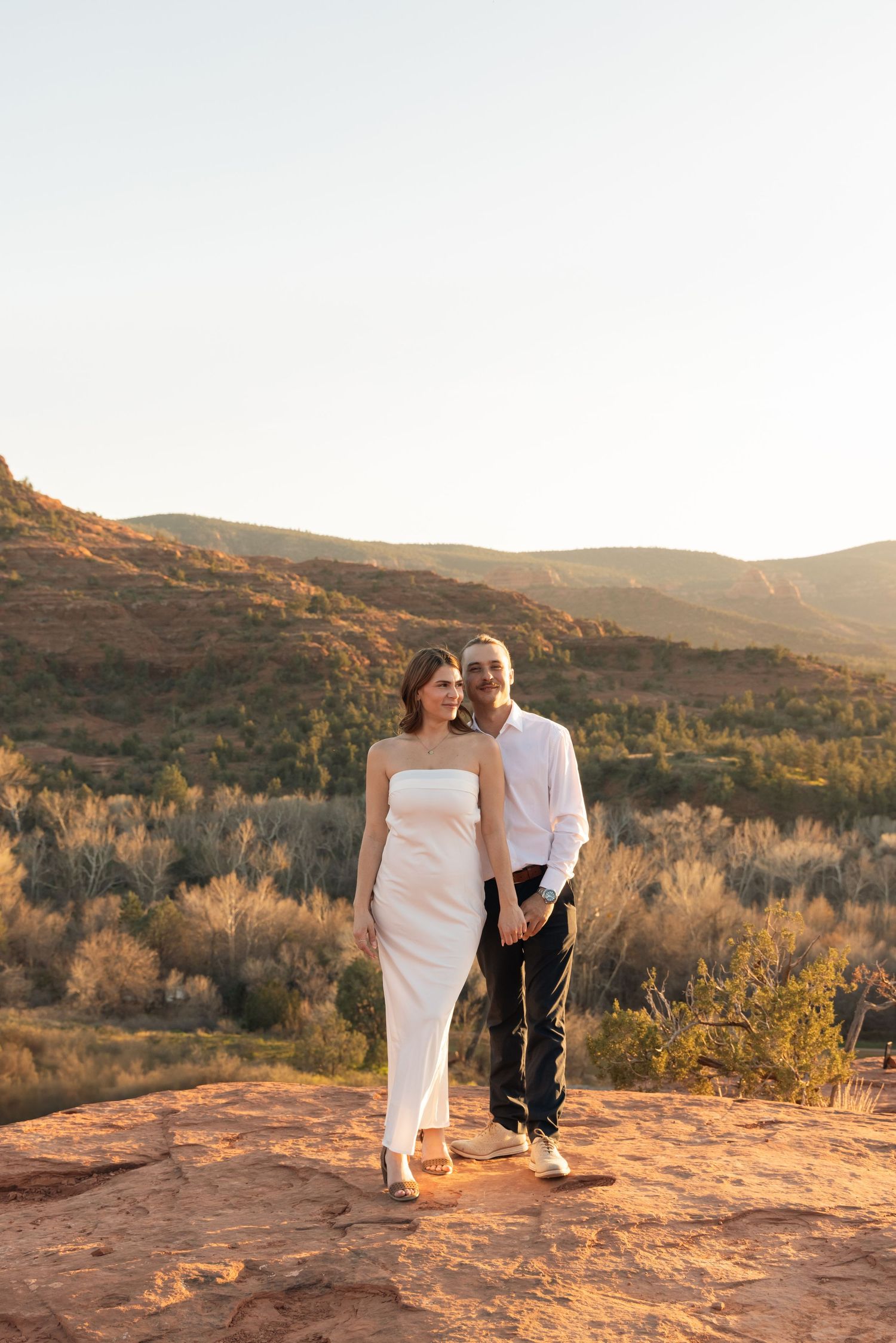 A couple stands together on a red rock cliff at sunset in the desert with mountains in the background.