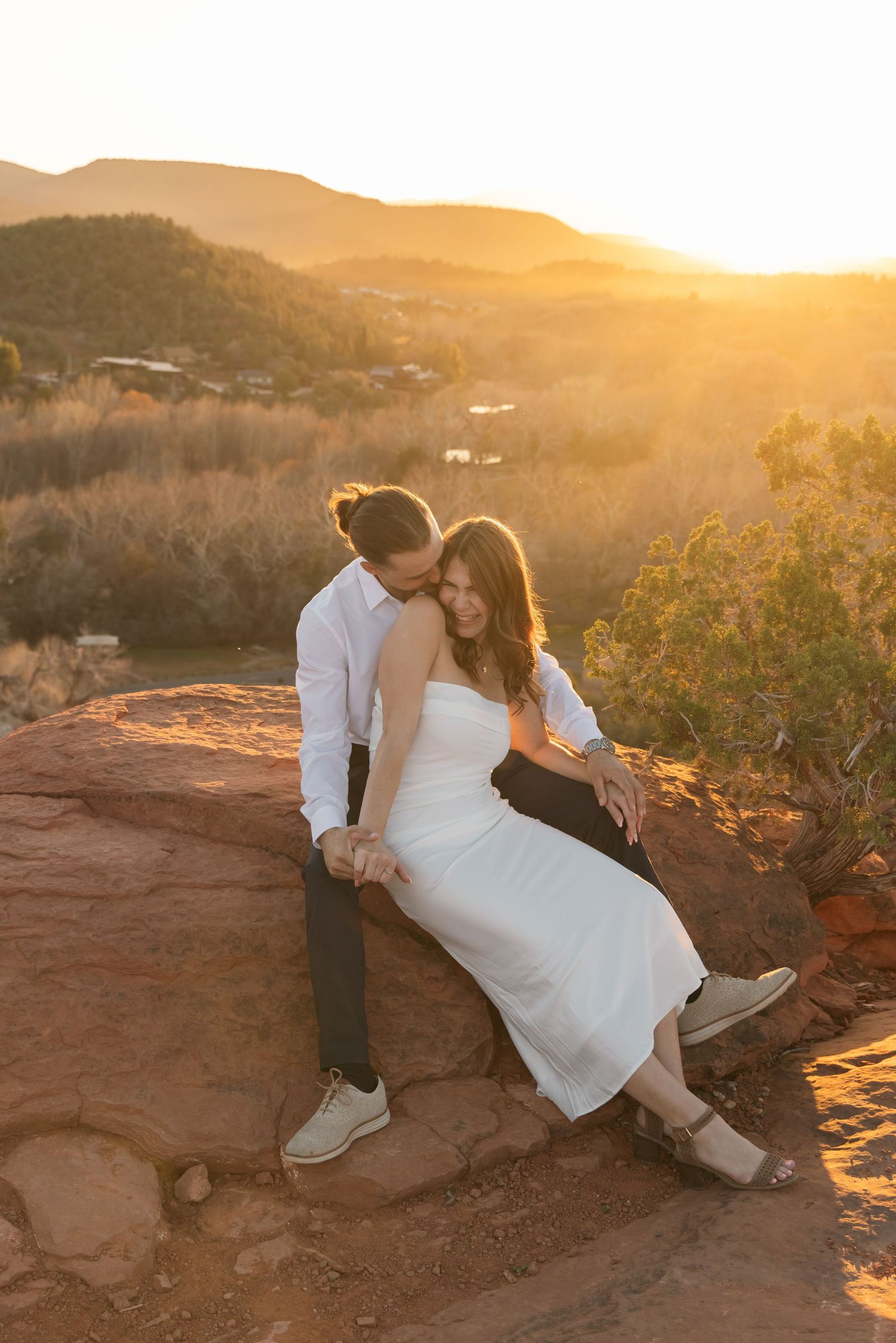 A couple in wedding attire embraces on red rocks during golden hour sunset in a desert landscape.
