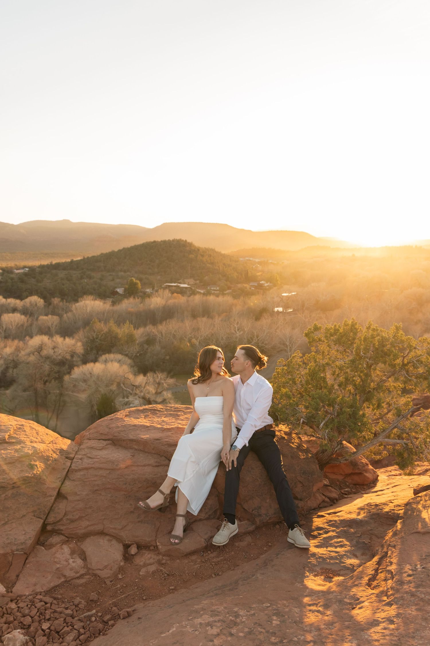 A couple sits together on red rocks overlooking desert mountains during golden hour sunset in Sedona.