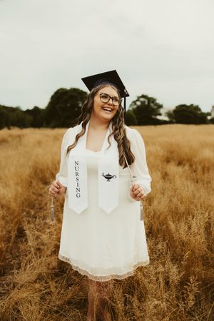 Proud nursing graduate smiles holding nursing stole in golden field wearing white dress and graduation cap.